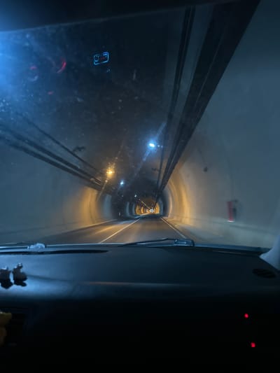 The image depicts an interior view from what appears to be a train or vehicle moving through a tunnel. The tunnel is circular in cross-section and is illuminated by intermittent lights. The walls of the tunnel are light in color, appearing either white or light gray. The center of the tunnel has two sets of parallel tracks. The perspective suggests the viewer is in the front of a train, and the tunnel extends into the distance, appearing smaller with each increment. In the vehicle, there is equipment attached to the walls. The area in the bottom right of the image is heavily shadowed, obscuring any details. The location information provided indicates this tunnel is likely within the city of Liaskovo, Bulgaria.