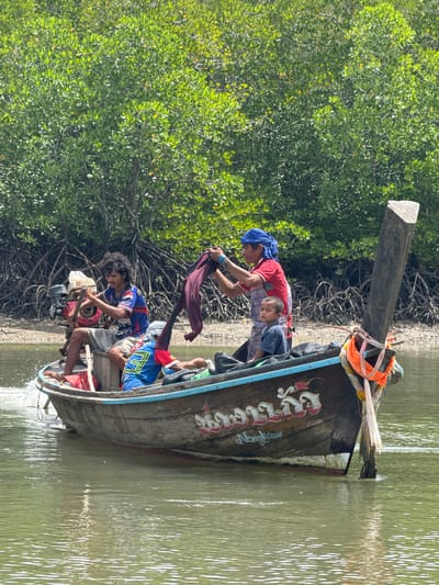 In Ban Hin Rom, Thailand, a wooden longtail boat navigates through calm, green water. The boat is occupied by three individuals: an adult man steering with a tiller and an outboard motor, another adult man holding a maroon cloth, and a young child sitting between them. The boat's hull is adorned with painted white and red lettering and a decorative design. In the background, a dense mangrove forest with its characteristic aerial roots lines the shore under a bright, sunny sky. The water reflects the surrounding foliage, creating a dappled surface.