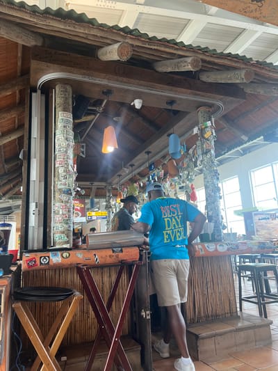 The image depicts a bar interior with distinctive decorations, including money attached to beams and walls. Patrons are present, one wearing a blue "BEST DAY EVER" t-shirt. Structural elements are made of wood, creating a rustic ambiance. The bar features writing and signatures, possibly customer inscriptions. Light fixtures illuminate the scene. A glimpse of the outside suggests the bar might be located in an airport or travel hub.
