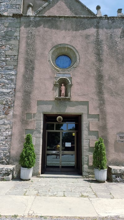The image shows the facade of a building, likely a church or sanctuary, with a rustic stone and plaster exterior. A large, rounded window with blue stained glass is positioned above a central entrance. The entrance itself is a set of double glass doors, reflecting the surroundings. Flanking the entrance are two potted evergreen trees. A small statue of a standing figure is housed in a niche above the doors. To the right of the entrance, a plaque is affixed to the wall, bearing text that reads "Santuario del Far" and "s. XV". The ground in front of the building is paved with stone slabs. The architectural style suggests a historical building. Based on the plaque, the location is identified as the Santuario del Far in Susqueda, Spain.