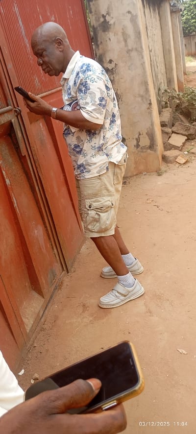 The scene depicts an outdoor setting in Abagana, Nigeria, captured on March 12, 2025, at 11:04 AM.
A male individual, appearing to be elderly with dark skin and a bald head, is standing and leaning against a large, weathered, reddish-orange corrugated metal gate on the left. He is dressed in a light-colored, short-sleeved collared shirt featuring blue floral patterns, khaki cargo shorts, white socks, and light-colored shoes with white velcro straps and a gold-colored accent. A silver-toned wristwatch is visible on his left wrist, and a dark band with a circular white emblem is on his upper left arm. The man is holding a black smartphone with both hands and is intently looking down at its screen.
In the lower foreground, a dark-skinned hand, presumably belonging to a second individual, is holding a dark-colored smartphone with a golden bezel; the thumb is positioned on the screen, indicating interaction.
The ground consists of reddish-brown dirt or earth, appearing unpaved and uneven. To the right of the standing man, a weathered concrete wall, discolored with dark patches, stands vertically. Beyond this wall, scattered pieces of debris are visible on the ground, and faint green foliage with distant structures can be observed in the background.