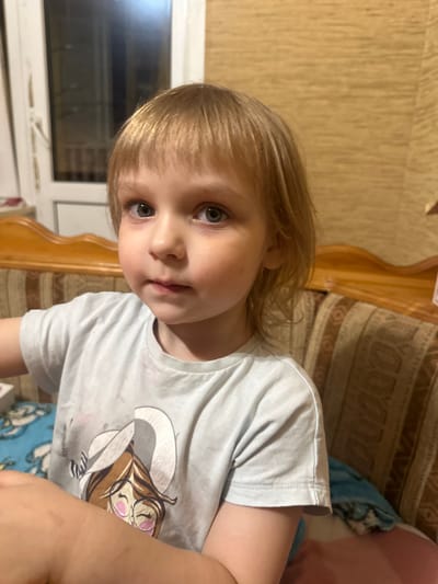 The image shows a young child with blonde hair and grey-green eyes sitting on a couch. The child is wearing a light blue t-shirt with a cartoon graphic of a girl wearing glasses. The couch has a patterned fabric and a wooden frame. In the background, there is a window and a wall with a textured wallpaper. The scene appears to be indoors, likely in a residential setting. The image is a close-up shot, focusing on the child's face and upper body. The lighting suggests it is evening or nighttime. The location is identified as Pokryshkino, Russia.