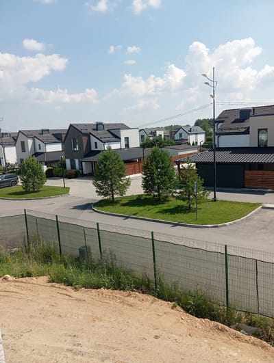 The image shows a street in the city of Terema, Russia, with several modern houses lining the road. The houses are two-story structures with dark-colored roofs and a mix of white and wood-toned exterior walls. Some of the houses have garages with dark-colored doors. The street is paved, and there are grassy areas with small trees and bushes in front of the houses. A green metal fence runs along the foreground, separating the street from a dirt embankment with wild grass. The sky is blue with scattered white clouds. There are no people or vehicles visible in the immediate scene. A street lamp is visible in the middle distance.