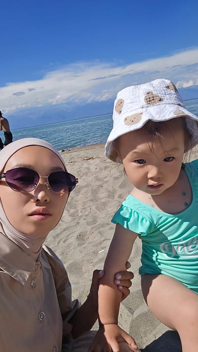 This is a selfie taken at Boz Beshik, Kyrgyzstan. The image features two subjects, an adult and a young child, on a sandy beach with the sea and sky in the background. The adult, partially visible in the upper left, is wearing a light-colored hijab and dark sunglasses. The adult's hand is holding the child's arm.
The child, in the foreground, is wearing a light blue swimsuit and a white bucket hat adorned with a teddy bear pattern. The child is looking towards the camera with a curious expression. The sea appears calm, with the horizon visible in the distance. The sky is bright blue with scattered white clouds, suggesting a sunny day, likely during daylight hours. The sandy texture of the beach is clearly visible. The overall mood of the image is peaceful and suggests a casual outing at the beach. There is no visible text in the image.