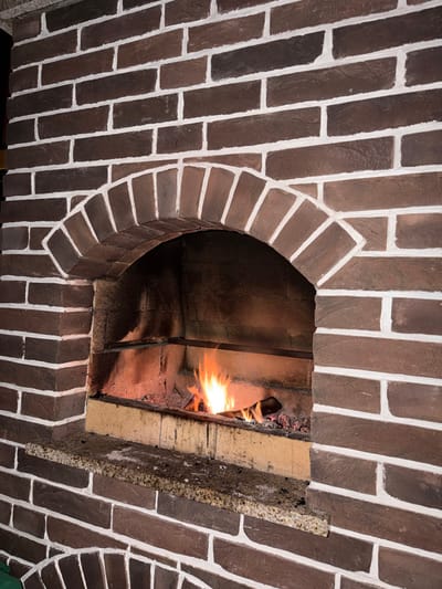 The image displays a close-up view of a static brick structure, identified as a fireplace or oven, located in Shchelkovo, Russia. The structure is constructed from dark brown bricks arranged in horizontal courses, meticulously highlighted by contrasting white mortar lines. A prominent arched opening is centered within the brickwork, leading into the firebox. Inside the firebox, a small fire is actively burning, characterized by bright orange and yellow flames emanating from several pieces of wood and glowing embers at the base. The interior walls of the firebox exhibit darkening and sooting from prior use. Directly beneath the firebox opening, a speckled, light brown granite slab functions as a hearth or mantelpiece. The brickwork continues above and surrounding the arched opening, indicating a robust construction. No individuals are present, and no specific actions or interactions are depicted.