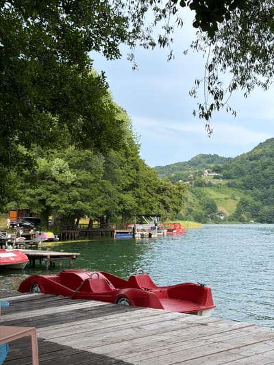 The image depicts a scenic view of a lake, with lush green hills in the background. Red pedal boats are docked on a wooden pier, ready for use. The water is calm and reflects the surrounding greenery. Trees line the shore, creating a natural and serene atmosphere. The sky is clear and bright, suggesting a pleasant day. The presence of pedal boats implies recreational activities, such as boating or leisurely paddling on the lake. The overall scene is tranquil and inviting, offering a peaceful escape into nature. The location is Ćusine, Bosnia and Herzegovina, a place known for its natural beauty. There are no visible people in the image, nor any discernible text.