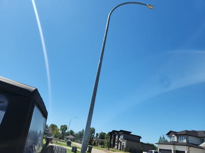 This image depicts a bright, sunny day in a suburban neighborhood, likely in White City, Canada. A large, curved street lamp dominates the right side of the frame, its metallic pole extending upwards against a clear blue sky. In the foreground, the corner of a dark-colored vehicle, possibly a jeep with a soft top, is visible on the left. The vehicle's tire is also visible.

The background reveals a street lined with modern houses. One house in particular, on the right, is a two-story dwelling with a dark roof, light-colored siding, and stone accents. Another house to its left features a similar architectural style. Green trees are scattered throughout the scene, and a few garbage bins are placed along the sidewalk. A streak of light, possibly from the sun, is visible in the sky. The overall impression is of a typical residential street on a pleasant summer day.
