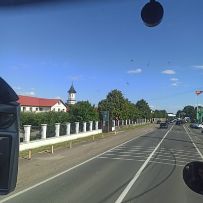 The image depicts a street scene in Rădăuți, Romania, on a clear, sunny day. The perspective is from inside a vehicle, likely a bus or car, looking forward and slightly to the right.

On the left side of the road, a white building with a red tiled roof is visible, adjacent to a church with a black, pointed steeple. A white fence with black ironwork runs along the property line, separating it from the road. Green trees and bushes line the area behind the fence.

The road itself is paved and has clear white lane markings, including a pedestrian crossing marked with thick white stripes. Several cars are driving on the road, with some parked on the right side. A prominent red and white MOL gas station sign is visible in the distance on the right. The sky is a bright blue with a few scattered white clouds. The overall impression is of a suburban or semi-urban environment.