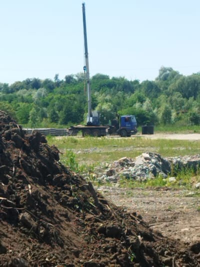 This image depicts an outdoor scene with a large blue crane truck parked on a dirt and gravel lot. The crane boom is extended upwards, reaching into a clear, light blue sky. In the foreground, a large pile of dark, disturbed earth and debris is visible. Behind the truck, a dense line of green trees and foliage forms the background, suggesting a rural or semi-developed area. To the right of the truck, a cylindrical dark object, possibly a manhole cover or a piece of construction material, rests on the ground. In the middle ground, stacks of rectangular concrete slabs are arranged on the ground, indicating construction or landscaping materials. The overall impression is a work-in-progress or a site where heavy machinery is actively used. The lighting suggests it is daytime, likely mid-morning or afternoon, with bright sunlight casting minimal shadows. No people are visible in the image. The scene appears to be in Rozdil, Ukraine, based on the provided location context, but there are no visible text elements to confirm this.
