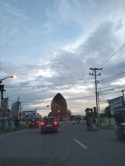 The image captures a street scene in Sigli, Indonesia, during twilight. The sky is overcast with diffused light, indicating either early morning or late evening. A prominent, tall, brown, dome-shaped structure, possibly a landmark or monument, stands on the right side of the road, adorned with the word "SIGLI" in large letters at its base. A traffic light with a red signal is visible near the structure.
The road in the foreground is asphalt, with a white line marking the lane. Several vehicles are present: a dark SUV in the center lane, its brake lights illuminated, and other cars and motorcycles further down the road. Some vehicles appear to be moving, while others are stopped at the traffic light.
On the left side of the road, there are buildings, streetlights, and utility poles with numerous wires crisscrossing the sky. A bright streetlight illuminates the immediate area on the left. On the right side of the road, there are more buildings, a green fence, and trees.
Several individuals are visible, mostly on motorcycles. One person is operating a motorcycle in the right lane, while others are further in the distance, moving along the road. The overall atmosphere is that of a typical urban street during a transitionary period of daylight.