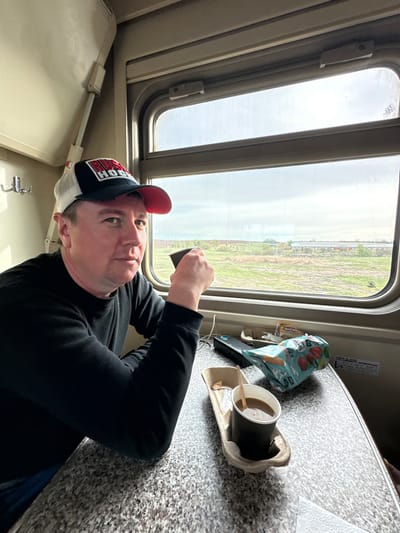 A man wearing a black long-sleeved shirt and a red and black baseball cap sits next to a train window. He is holding a small black object in his raised hand. A cup of coffee in a paper holder sits on a speckled table near a bag of potato chips. The window reveals a landscape of green fields and distant buildings. The image was likely taken on a train in Уральский, Russia.