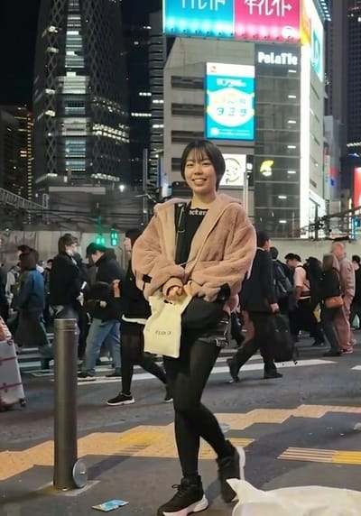 The image depicts a crowded nighttime street scene, centered on a young East Asian woman smiling directly at the camera. She stands in the foreground, wearing a light pink faux-fur hooded jacket over a black top with white lettering, black shorts, black tights, and black and white athletic shoes. She holds a small white tote bag with "ELOC F" text in her left hand, and a black cross-body bag is visible. To her left, a dark grey bollard is visible on the sidewalk. On the ground near the bollard, a piece of light blue refuse is present.

In the midground and background, numerous pedestrians, primarily East Asian adults, walk in various directions across a street marked with white crosswalk lines and yellow tactile paving. They are dressed in typical urban winter attire.

The background features multiple tall buildings illuminated with numerous windows, indicative of a dense urban environment. Prominently visible are large, brightly lit neon signs in Japanese. One large sign features horizontal blue and pink sections with Japanese text. Below it, a vertical sign displays "Polatte" and additional Japanese text with numerical figures (e.g., "329"). To the left, a distinctive skyscraper with a lattice-like exterior, identifiable as the Mode Gakuen Cocoon Tower, rises into the night sky.

Based on the presence of the Mode Gakuen Cocoon Tower and the general urban architecture and neon signage, the location is **Shinjuku, Tokyo, Japan**, not Gifu.