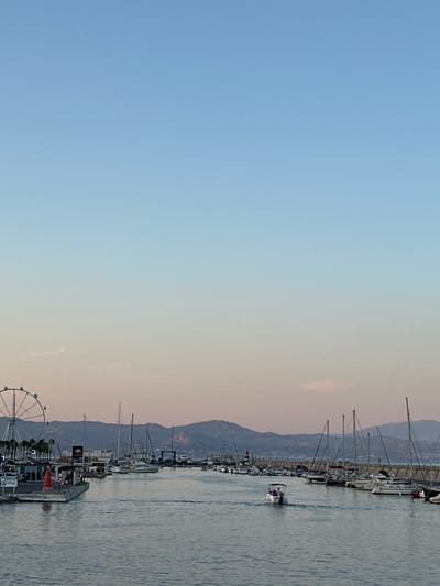 The image depicts a harbor scene in Arroyo De La Miel-Benalmadena Costa, Spain, during what appears to be either sunrise or sunset, judging by the gradient of the sky. In the foreground, the calm blue water of the harbor reflects the soft light. A small white boat with two individuals is navigating through the water, heading away from the viewer. Along the left side of the harbor, a floating platform is visible with a red lighthouse-like structure, palm trees, and a large Ferris wheel in the background. Numerous sailboats and yachts are moored along the right side of the harbor, creating a dense line of vessels. A stone breakwater extends into the harbor, with more boats docked behind it. In the distance, a range of hills or mountains forms the horizon, silhouetted against the pastel-colored sky, which transitions from a light blue at the top to a pale pinkish hue closer to the horizon.