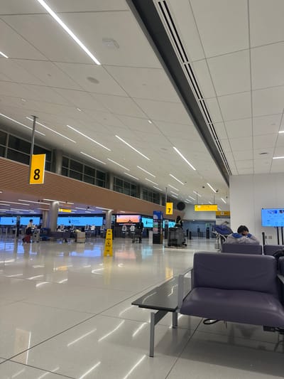 The image depicts an airport terminal concourse, specifically the terminal area with gates 7 and 8 in Denver, United States of America. The scene shows rows of empty purple seating on the right, a glossy tiled floor reflecting overhead lights, and people walking by signs for Bag drop, Check-in 1-6, and caution signs. The overhead signage indicates gate numbers. People are generally seen walking past various service desks.