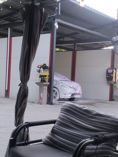 A car is being washed with foam at a self-service car wash in Șoldănești, Moldova. The car is covered in pinkish-white foam, and its front is visible. In the foreground, a dark grey curtain hangs from a structure, tied with a rope. To the right of the curtain, a yellow and grey cleaning station with soap dispensers is visible. A payment machine with a digital display showing "154.9 SPUMA" is mounted on a post to the right of the car wash bay. The scene appears to be outdoors, likely during the day, as indicated by the lighting. The overall atmosphere is functional, focusing on the car cleaning activity.