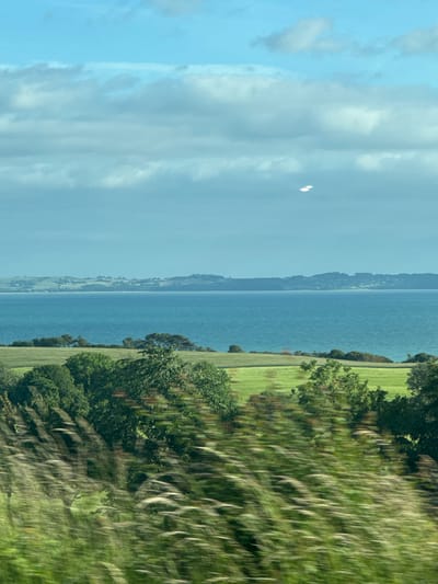 The image depicts a scenic view of a coastal landscape. In the foreground, tall grasses are blurred due to motion, suggesting the image was taken from a moving vehicle. Behind the grasses, a vibrant green field stretches out, dotted with various trees and shrubs. Beyond the field, a vast expanse of blue water, likely an ocean or bay, is visible. In the distance, rolling hills covered in greenery and scattered with buildings form the horizon. The sky above is a pale blue, with scattered white clouds, and two bright, elongated shapes are visible, possibly lens flares or reflections. The scene conveys a sense of natural beauty and tranquility. The location is not identified, and no people or specific actions are depicted.