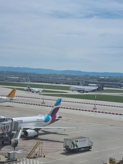 The image shows an aerial view of a section of Stuttgart Airport in Echterdingen, Germany. Several airplanes are visible, including a Eurowings plane at the foreground dock, a Pegasus Airlines plane at the bottom of the shot, a Kuehne + Nagel cargo plane in the mid-ground, and another unidentified passenger plane at the top center. There is a gate gourmet truck next to the Eurowings plane. The cityscape is visible beyond the airport grounds, with mountains in the distance, and a clear blue sky.