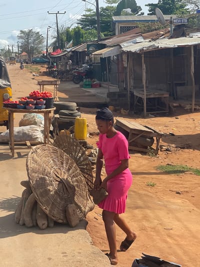 A young woman in a pink dress stands in a marketplace in Ikoga, Nigeria. She leans over a large, round wicker basket filled with yams. Next to her is a wooden cart laden with produce including tomatoes, bags of goods, tires, and a yellow container. In the background are shops with corrugated metal roofs. There are motorbikes parked and a street sign that reads "WEVI STR." The environment is sunny and dusty, with dirt roads and sparse vegetation.