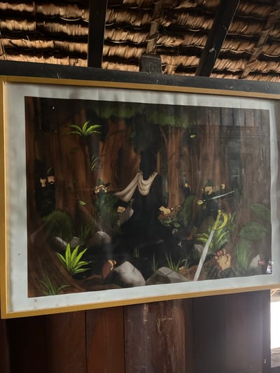 This image depicts a framed painting of a group of figures in a forest setting. The painting has a thick, light-colored border and is hung against a background of wooden planks, with a thatched roof visible in the upper portion of the frame. The painting itself shows several stylized figures, some holding weapons such as swords. The central figure is wearing dark clothing and a white sash. The environment appears to be a dense forest with trees and foliage. The painting seems to be displayed indoors, possibly in a traditional-style building, given the wooden walls and thatched roof. The location is specified as Mesjidpirak, Indonesia.