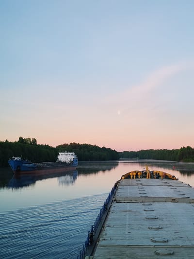 The image captures a serene river scene during twilight, likely at either sunrise or sunset, in Volokov Most, Russia. The perspective is from the deck of a cargo vessel, with its front deck, featuring numerous hatch covers and yellow machinery, visible on the right side of the frame. Blue railings line the left edge of this vessel's deck.
In the midground, another large cargo ship, painted blue with a white superstructure, navigates the calm, reflective waters of the river, moving in the opposite direction. Its clear reflection is visible in the water, along with the gentle wake from the foreground vessel.
The river is bordered by dense green forests on both banks, suggesting a natural or semi-natural waterway. A subtle red buoy can be seen in the distance on the right bank. The sky transitions from a soft pale blue at the top to a warm pink and orange near the horizon, illuminating a distinct, bright white moon positioned in the upper right quadrant. No people or discernible text are visible in the tranquil scene, which focuses on the peaceful passage of vessels through a natural landscape.