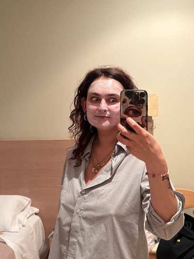 A selfie taken indoors shows a woman with dark curly hair and a white facial mask. She holds a black smartphone with a graphic eye design on the case. She is wearing a gray button-down shirt and gold jewelry. A pink beaded bracelet is visible on her left wrist. A bed with white linens and a headboard are behind her. The location is likely an interior space in Puçol, Spain.