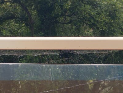 The image shows a close-up of what appears to be a railing or ledge with a blurred background of trees and foliage. The top portion of the image is a solid, light-colored surface, possibly painted wood or metal, with a few small water droplets visible. Below this is a textured layer that resembles a spiderweb, intricate and delicate, catching light and appearing somewhat metallic in places. This spiderweb layer is overlaid on a darker, mottled green and black surface, which might be vegetation or a patterned material. Beneath this is a dark, reflective surface, likely glass or polished stone, showing a distorted reflection of the trees and sky. The overall scene is out of focus, with the focus being on the foreground elements. There are no people, animals, or specific landmarks identifiable in this image. It is impossible to determine the location in Arucas, Spain, based on the provided visual information.