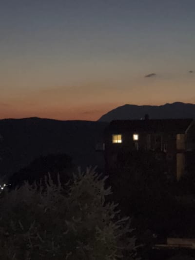 The image depicts a dimly lit scene at dusk, with a gradient of colors in the sky transitioning from a muted orange and pink near the horizon to a deep blue and gray above. Silhouetted against the sky are distant mountain ranges. In the mid-ground, a dark building with illuminated windows stands on a dark hillside. The windows emit a warm, yellow light, suggesting the interior is lit. In the foreground, the dark silhouettes of foliage, possibly bushes or trees, are visible, with some lighter, feathery-looking branches or leaves catching faint light. The overall impression is a quiet, residential setting nestled in a hilly or mountainous landscape as night begins to fall. The location is identified as Svinjišta, Montenegro.