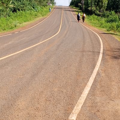 The image shows a paved road in Ihururu, Kenya, with a yellow centerline and white edge lines. The road curves slightly uphill. Three people are walking along the right side of the road, while one person is walking on the left side further ahead. Green vegetation lines both sides of the road. Utility poles are visible in the distance. The road surface is textured and appears worn.