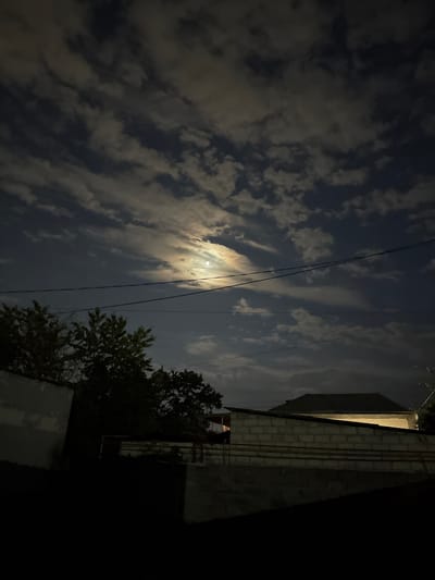 This media file depicts a nighttime scene in Nogir, Russia. The primary subjects are the moon, partially obscured by clouds, and the night sky. The scene is set outdoors, with elements of a building visible on the right side of the frame, constructed from light-colored bricks. Power lines are strung across the sky, contributing to the urban or suburban setting. A tree with dark foliage is also present, partially silhouetted against the sky.

The image captures a moment of natural beauty at night, with the moon illuminating the clouds. The overall atmosphere is tranquil and serene. The time of day is clearly night, indicated by the darkness of the sky and the presence of the moon. No specific activity or event is taking place, beyond the passive observation of the night sky. There is no visible text in the image. The location context of Nogir, Russia, provides geographical information about where the photo was taken.
