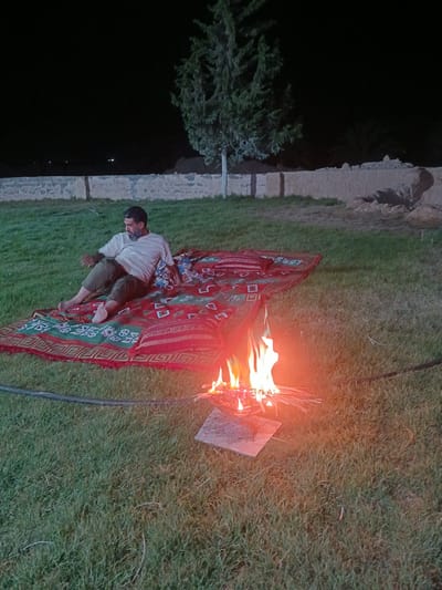 An adult male with a beard, wearing a light-colored t-shirt and dark shorts, is sitting on a red and green patterned blanket spread on grass. He is reclining and looking to his left. Next to him on the blanket is a bundle of fabric, possibly pillows. In the foreground, a small campfire burns on a square stone. The scene is outdoors at night, with a dark sky and a dimly lit stone wall in the background. A tree is visible behind the wall. The overall setting appears to be a park or outdoor recreational area in Hun, Libya.