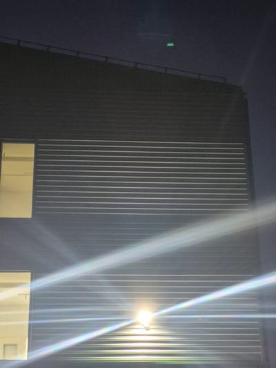 This image depicts the exterior of a modern building at night in Pabradė, Lithuania. The building's facade is constructed of dark, horizontally ribbed siding, creating a textured appearance. A railing runs along the top edge of the building. There are at least two large, rectangular windows visible; one on the left side of the image, brightly lit from within, and another lower window, also illuminated. A prominent security light fixture is mounted on the building, emitting a strong, bright light that causes significant lens flare, creating diagonal streaks of rainbow-colored light across the image. In the dark night sky above the building, a small, rectangular, green light is faintly visible. The overall scene suggests an industrial or commercial setting illuminated by artificial light against a dark, nocturnal sky.
