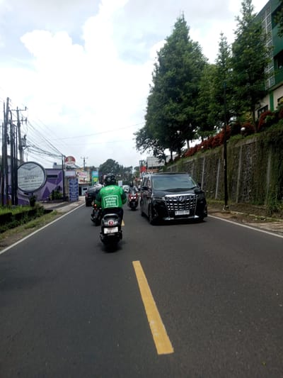 The image depicts a street scene in Ban Phouviengnoy, Laos. A black Toyota Alphard is driving on the right side of the road, while a motorcycle with a Grab driver in a green jacket is in the lane to the left. The driver's jacket displays the Grab logo and the text "Isi Pulsa Murah di Grab." A yellow lane divider is visible on the road. On the left side of the street, there are buildings, including one with a sign that reads "Signature Hotel Wedding Hall & Meeting Room" and "GINGARAJA". Overhead power lines are visible. Trees and a wall are on the right side of the road, with some red flowers planted on top of the wall. The sky is overcast with cloudy conditions.