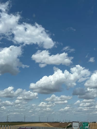 The image depicts a wide view of a bright, cloudy sky over a rural landscape, likely near Cruceni, Romania. Cumulus clouds, varying in size and density, dominate the upper half of the frame, against a backdrop of deep blue sky. Below the clouds, a flat, open field is visible, stretching towards a distant treeline. Several tall electricity pylons are scattered across the landscape, indicative of power transmission infrastructure. In the foreground, a highway is present, with a white truck bearing the "RAVITEX" logo and a green directional signpost indicating routes to "Szeged," "Varsa," and "Arad." A small section of a light-colored barrier runs along the left edge of the highway, and a glimpse of a dark-colored car is visible on the road. The scene suggests a sunny day, possibly during daylight hours, with clear visibility.