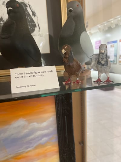 The image depicts an indoor museum display featuring various pigeon figures. In the foreground, on a clear glass shelf, are two small, hand-crafted pigeon figures: one brown on the left and one light gray on the right. Both small figures feature red feet and silver leg bands. A white rectangular sign on the glass shelf states, "These 2 small figures are made out of instant potatoes. Donated by Ed Pointer." Behind the glass shelf, two larger pigeon sculptures are visible: a solid black one on the left facing left, and a gray and white one on the right facing right. A framed black and white artwork is partially visible behind the black figure. Below the glass shelf, a portion of a framed painting, depicting a sky with orange and purple clouds, is visible. The background shows blurred elements consistent with a museum or gallery setting, including informational posters on walls. The scene is located at the American Racing Pigeon Union Hall of Fame Museum in Oklahoma City, Oklahoma, United States. No people are visible, and the scene is static.