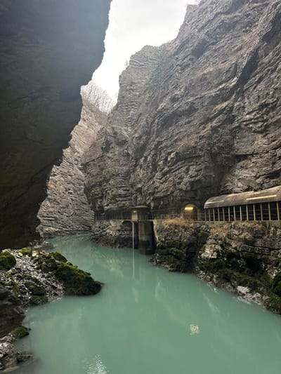 The image displays a deep, narrow gorge in Pellafol, France. A calm, light blue-green river flows between towering, steep-sided rock cliffs of layered grey stone. The sky above is overcast. On the left, a dark rock face dominates the foreground, with moss-covered rocks and debris at the water's edge. Along the right cliff face, a series of human-engineered structures are visible: a multi-level concrete platform with a vertical pipe extending into the water, a narrow path or walkway, and a warmly illuminated tunnel entrance. A wooden-slatted covered structure with a sloped roof extends outward from the tunnel mouth. Sparse vegetation, including bare trees and brush, clings to the upper parts of the cliffs. Overhead, power lines are present, traversing the gorge against the sky. No individuals, actions, or interactions are observed.