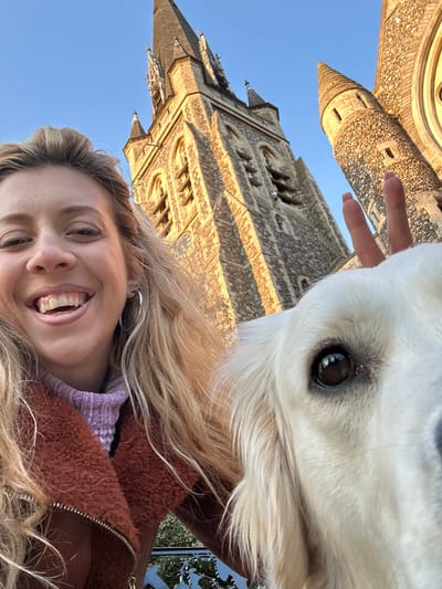 An outdoor, eye-level photograph depicts a light-skinned female and a light-colored dog posing in front of St. Thomas of Canterbury Church in Brentwood, Essex, United Kingdom.

The female, positioned on the left, has long, wavy blonde hair and is smiling broadly, showing her teeth. She wears silver hoop earrings, a lilac ribbed turtleneck, and a brown, textured jacket with a visible zipper. Her left hand, with orange-pink painted fingernails, is partially visible behind the dog's head, forming a two-finger upward gesture.

A light-colored dog, likely a Golden Retriever, occupies the right side of the frame, with its head and chest visible. Its right eye is prominent, dark, and reflects light, indicating it is looking towards the camera.

In the background, St. Thomas of Canterbury Church is prominently featured. The church exhibits a tall, pointed spire surmounting a square tower constructed from light-colored stone, displaying multiple windows and architectural details. To the right of the main tower, a smaller, conical turretted structure is visible. The church is bathed in warm, directional sunlight, contrasting with the clear blue sky above. A decorative metal railing is partially visible at the bottom center of the frame.