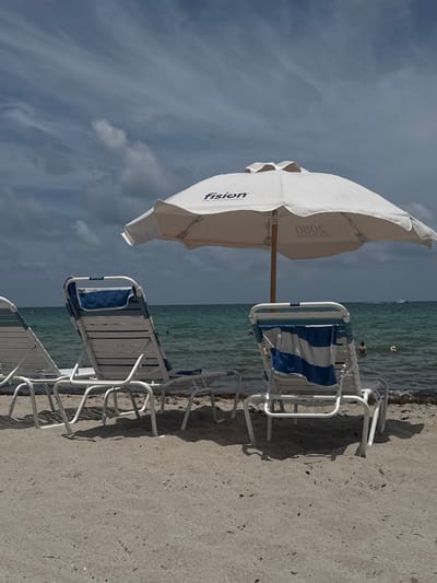 The image captures a beach scene in Hallandale Beach, United States. The foreground is composed of light-colored sand, upon which three white-framed beach loungers with blue and white striped fabric are positioned, facing the ocean. The leftmost lounger is partially visible. The central lounger has a solid blue towel draped over its upper back, while the rightmost lounger holds a blue and white striped towel. A large white beach umbrella stands upright, slightly right of the frame's center. The word "fision" is printed in blue on one side of the umbrella, and "080S" is visible in a mirrored orientation on its opposite underside.
Beyond the sand, the clear turquoise-green ocean water extends to the horizon. A narrow strip of dark organic material, consistent with seaweed, is present at the waterline. Two individuals are visible in the water; one is partially submerged near the center-right, and a second, less distinct figure is further out. In the distant background, a white powerboat traverses the water. The sky is significantly overcast with a blend of light and dark gray clouds.
