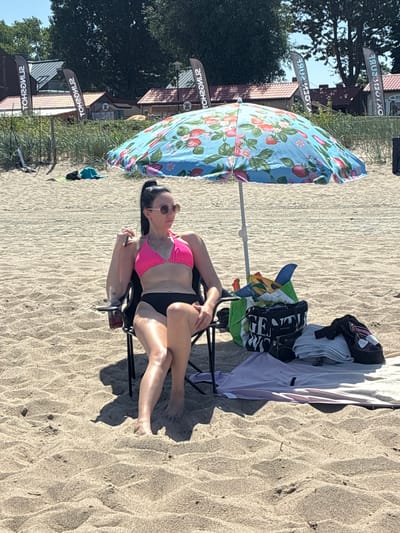 The image depicts a sunny day on the sandy beach of Rewa, Poland. A woman in a pink bikini and black bottoms is seated in a black camping chair, partially shaded by a colorful umbrella decorated with strawberries. She is wearing sunglasses and holding a drink in her right hand. Beside her, on the sand, are beach essentials including a green bag filled with items, a black tote bag with the word "GENETIC" visible, folded towels, and other clothing. In the background, several buildings with thatched roofs and flags with Polish text are visible, suggesting a beachside establishment or service area. The overall scene conveys a relaxed atmosphere of a day at the beach.