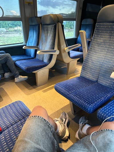 This is a first-person perspective image taken from inside a train carriage. The seats are upholstered in blue fabric with a white geometric pattern. In the foreground, the legs and feet of two individuals are visible. One person is wearing grey jeans and white sneakers, while the other is wearing similar grey jeans and grey athletic shoes. The floor of the carriage is a light tan color. Through the windows, greenery and a cloudy sky are visible, suggesting the train is in motion or stationary in a rural or suburban area. The specific location within the city of Alès, France, cannot be determined from the visual information in the image.