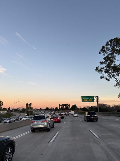 A vertical shot depicts a congested highway at sunset. Traffic is dense, with multiple vehicles visible. Above, a clear sky transitions from blue to orange hues. A road sign for "Bellflower Blvd" is seen. In the top left, a tree branch adds natural detail. The image is tilted, presenting an unusual perspective. The setting suggests an urban area experiencing heavy traffic during twilight.