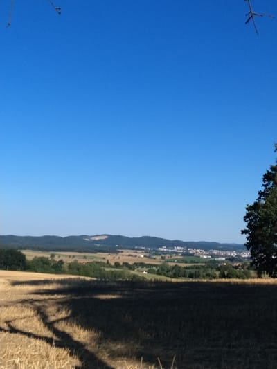 The image shows a rural landscape under a clear blue sky. In the foreground, a harvested field of straw casts long shadows. A large tree stands on the right side of the frame. In the middle ground, rolling hills covered in green trees and fields are visible. In the distance, a town with buildings and infrastructure stretches across the horizon, nestled amongst the hills. The overall scene is one of natural beauty and rural settlement. The specific location of Čížová, Czechia is not discernible from the image itself.