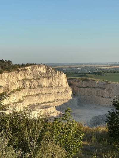 The image depicts a quarry in the city of Verbka, Ukraine. The quarry features steep, layered cliffs made of light-colored stone, likely limestone, showing evidence of excavation. Vegetation, including trees and shrubs, grows along the rim of the quarry and in the foreground. In the distance, a rural landscape unfolds with fields, scattered buildings, and trees under a clear, pale blue sky. The lighting suggests it is late afternoon or early evening, casting shadows across the quarry walls. There are no people or animals visible in the image.