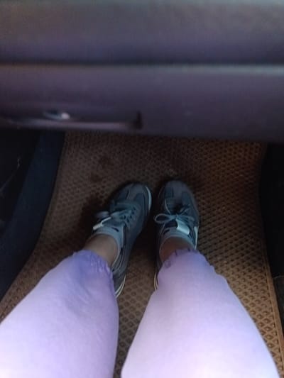 The image shows a first-person perspective from inside a vehicle. The lower half of a person wearing lavender-colored leggings and gray sneakers is visible. The feet are resting on a brown patterned car floor mat. The dashboard of the vehicle is visible above the feet. The location of the image is not specified, so it is impossible to determine the place and city Khyrkasy, Russia.