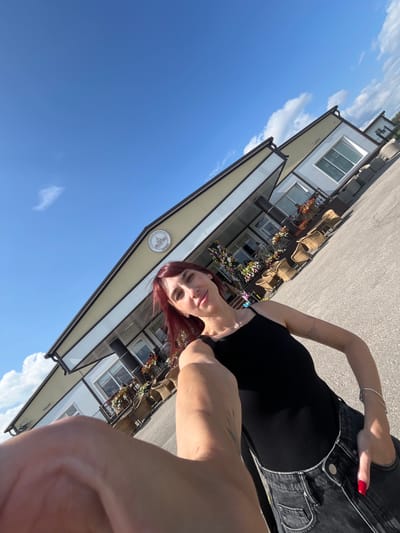 A woman with red hair and fair skin takes a selfie outdoors in front of a building. She is wearing a black tank top and black jeans. The building appears to be a commercial establishment, possibly a cafe or shop, with outdoor seating and potted plants. The sky is blue with scattered clouds. The image is slightly tilted. The location is identified as Akhlebaikha (Kovriginskii S/S), Russia.