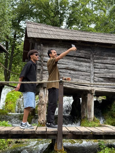 The image shows two young men standing on a wooden walkway over a stream. The man on the left is wearing a black short-sleeved shirt, light blue denim shorts, and sneakers. He has curly hair and is wearing sunglasses. The man on the right is wearing a tan t-shirt and black pants. He is holding a smartphone and appears to be taking a picture or video.
The setting is a natural environment with lush green trees and vegetation surrounding a wooden structure that looks like an old mill. Water from the stream is cascading down rocks, creating a picturesque scene. The wooden walkway is constructed from weathered planks, and a thick rope runs along its side, likely for support.
The time of day appears to be daytime, with natural lighting filtering through the trees. The weather seems pleasant, given the outdoor activity. The overall mood of the image is casual and adventurous, with the men enjoying their time in nature.
There is no visible text in the image. The location context is Prisoje, Bosnia and Herzegovina, which suggests a rural or semi-rural area with natural water features and traditional structures.