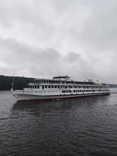 A white river cruise ship named "ВОЛГА ДРИМ" is traveling on a body of water. The ship has multiple decks with rows of windows and balconies. It is a cloudy day, and the sky is overcast. The ship is passing by a wooded shoreline. The water is dark and choppy, with some ripples and small waves. The overall impression is a serene and somewhat somber atmosphere. The ship appears to be moving from left to right in the frame. The name of the ship is clearly visible on its side.