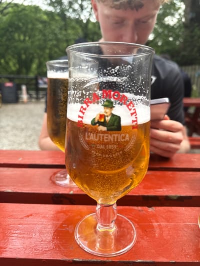 The image shows a person, presumably a young man, sitting at an outdoor table with a glass of Birra Moretti beer in his right hand. He is using his left hand to hold a smartphone, possibly to take a selfie or to message someone. The table is red and wooden. Another glass of beer is out of focus in the background. The table is on a patio surrounded by greenery. This scene is in Hayfield, United Kingdom, which is located at latitude 53.377 and longitude -1.935. The person appears to be enjoying a drink and using their phone.