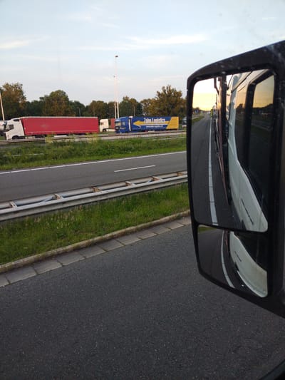 The image shows a view from a truck's side mirror, looking out onto a highway with other trucks. In the distance, a red semi-trailer truck and a blue semi-trailer truck with "Zahn Logistics" written on it are visible. The sky is clear with some clouds, and the sun appears to be setting, casting a warm glow on the scene. The highway is paved, with a metal guardrail separating it from the grassy shoulder. The mirror reflects the side of the truck from which the photo is taken, as well as the highway ahead. The image does not provide any specific information or landmarks to determine the location within the city of Aachen, Germany.