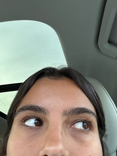 A close-up image depicts the upper portion of a human face, from the forehead to the bridge of the nose, situated within a vehicle interior. The individual has dark brown hair, prominent dark brown eyebrows, and dark brown eyes. The eyes are rolled upwards and slightly outwards, with the left eye gazing towards the upper-left and the right eye towards the upper-right. Above the subject's head, a light grey vehicle headliner is visible, along with a sunroof or moonroof opening that reveals an expanse of bright, light-colored sky. A black trim element is visible on the left side of the sunroof opening. On the upper right, a portion of a light grey sun visor and a light-colored car seat headrest are discernible. The precise location within Plainsboro Township, United States, is not visually discernible from the image content.