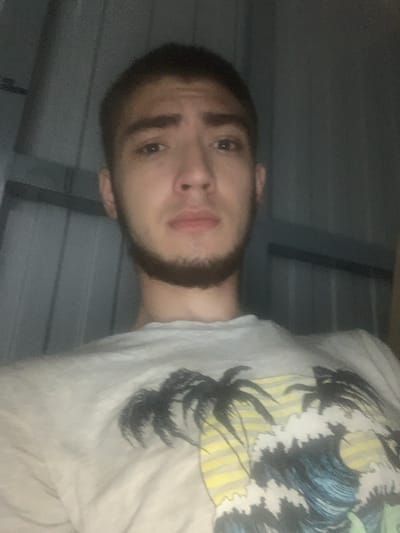 A young man with short, dark hair and a beard is shown from the chest up, looking directly at the camera. He is wearing a light-colored t-shirt with a graphic design featuring palm trees and waves. The background appears to be a wall with vertical lines. The lighting is dim, creating a grainy texture in the image. The image was taken in Kokhma, Russia.